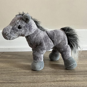 Op de afbeelding: Een grijs pluche paardenspeelgoed met een lange, vloeiende manen en staart. Het paard heeft een zachte, knuffelige textuur en is perfect voor kinderen om mee te spelen.