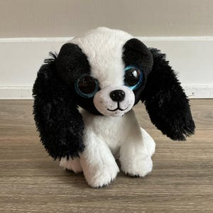 以下が含まれることがあります： 黒と白のぬいぐるみ犬。 大きなキラキラ光る青い目と長い黒い垂れ耳があります。 おもちゃの犬は小さな黒い鼻をしており、木製の表面に座っています。