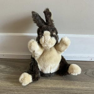 Può includere: Un coniglio di peluche marrone e bianco con orecchie lunghe e coda soffice. Il coniglio è seduto su un pavimento di legno.