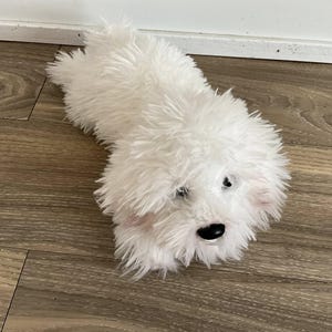 Pode incluir: Um brinquedo de pelúcia branco em forma de cão, com um pelo macio e texturizado. O brinquedo tem olhos pretos e um nariz preto, com detalhes rosa nas orelhas. O cão está deitado numa superfície de madeira.