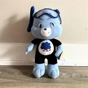 May include: A blue plush Care Bear wearing a black shirt and shorts with a cloud and heart design. The bear has a grumpy expression and is wearing a blue swim cap and goggles. A tag is attached to the bear's foot.