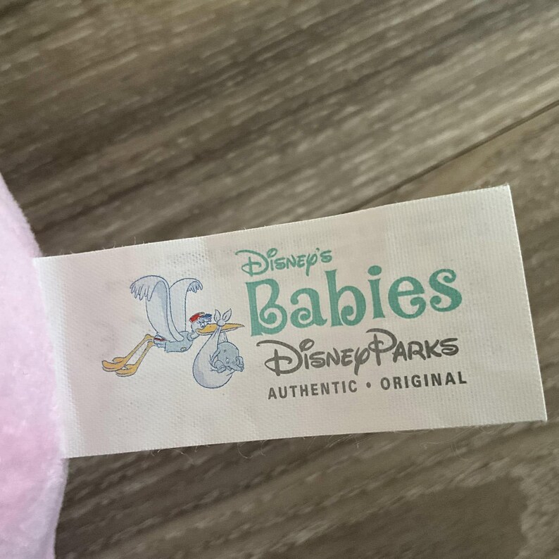 Pu&ograve; includere: Un'etichetta bianca con il testo "Disney's Babies Disney Parks AUTHENTIC &bull; ORIGINAL". L'etichetta presenta una cicogna che trasporta un bambino in un fagotto. L'etichetta &egrave; su un peluche rosa.