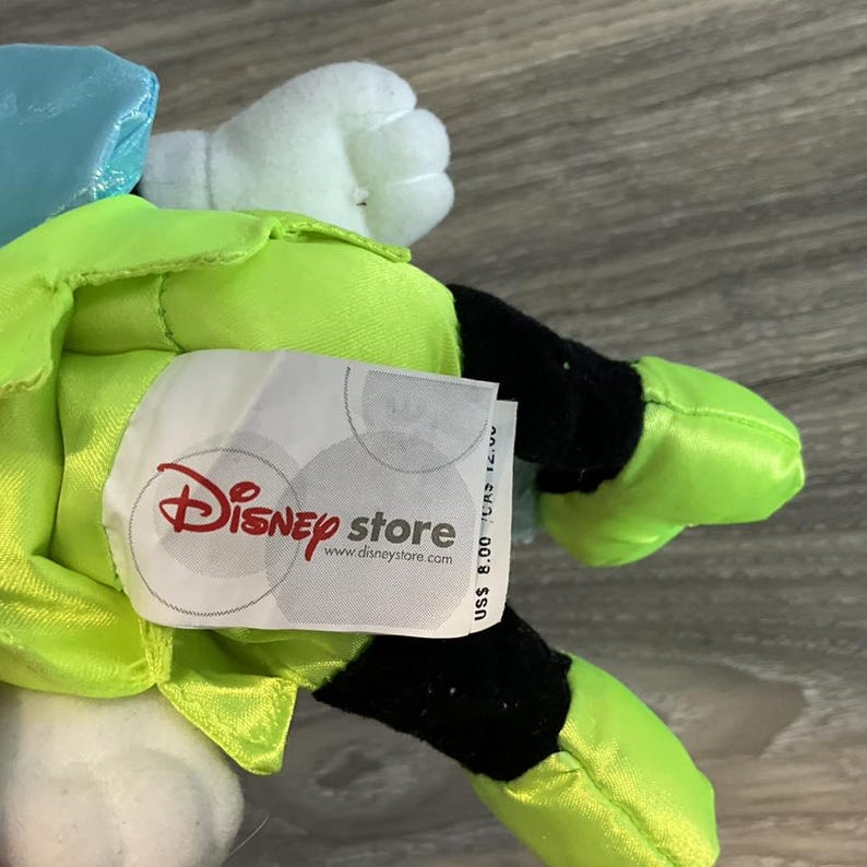 May include: A plush toy with a lime green body, black details, and white gloves. The toy has a blue head and a tag with the Disney Store logo and website address.