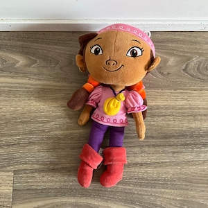 Può includere: Un giocattolo di peluche di una bambina pirata con i capelli castani, che indossa una camicia rosa con una borsa gialla, pantaloni viola e stivali rossi. Indossa una fascia rosa con cuciture bianche.