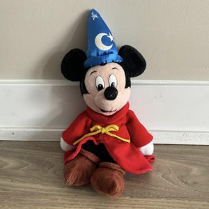 May include: A plush Mickey Mouse toy dressed as a sorcerer. Mickey wears a blue wizard hat with a white crescent moon and stars, a red robe with a yellow tie, and brown boots. The toy is sitting on a wooden floor.