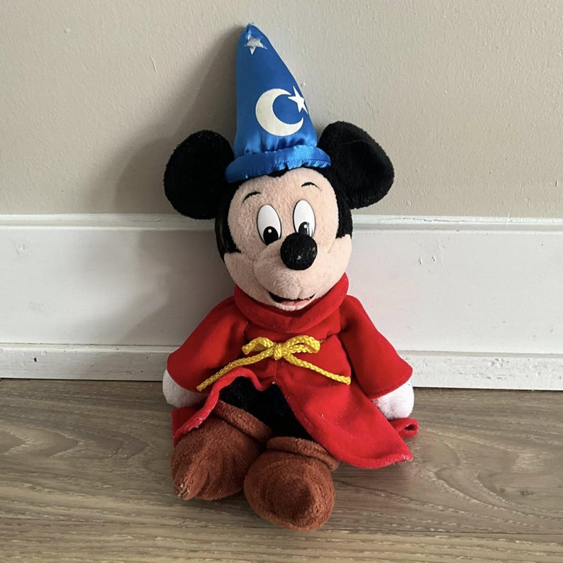 May include: A plush Mickey Mouse toy dressed as a sorcerer. Mickey wears a blue wizard hat with a white crescent moon and stars, a red robe with a yellow tie, and brown boots. The toy is sitting on a wooden floor.