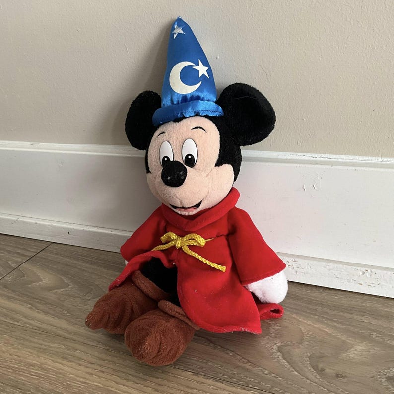 May include: A plush Mickey Mouse toy dressed as a sorcerer. Mickey wears a blue hat with a crescent moon and star, a red robe with a gold tie, and brown shoes. The toy is black, white, red, blue, and brown.