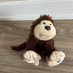 May include: A soft toy monkey with brown fur and a beige face, hands, and feet. The monkey has a black nose and a stitched smile. The feet have a colorful logo. The toy is on a wooden surface.