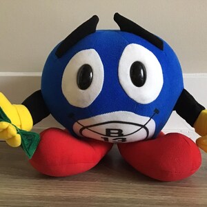 May include: Blue plush toy with black eyebrows, large white eyes, and a red mouth. The toy has yellow hands and feet and a green object in its left hand. The toy has a blue circle on its stomach with the number "B13" written on it.
