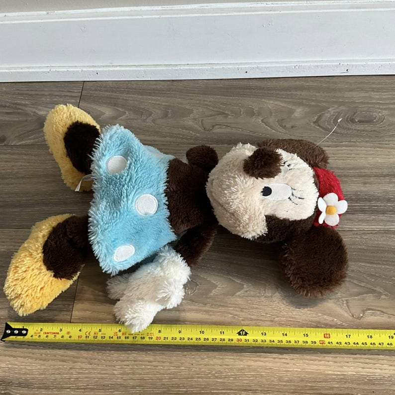 May include: A plush toy dog with brown, tan, and white fur, wearing a light blue outfit with white polka dots. The dog has a red bow and a white flower on its head. The toy measures approximately 18 inches in length.