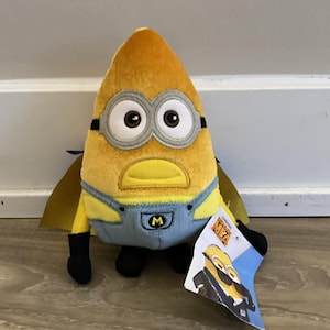 May include: A plush yellow Minion toy with large eyes, wearing blue overalls and a gold cape. The toy has black feet and a tag attached. The tag features a Minion in a suit. The Minion has a yellow body and is wearing silver goggles.