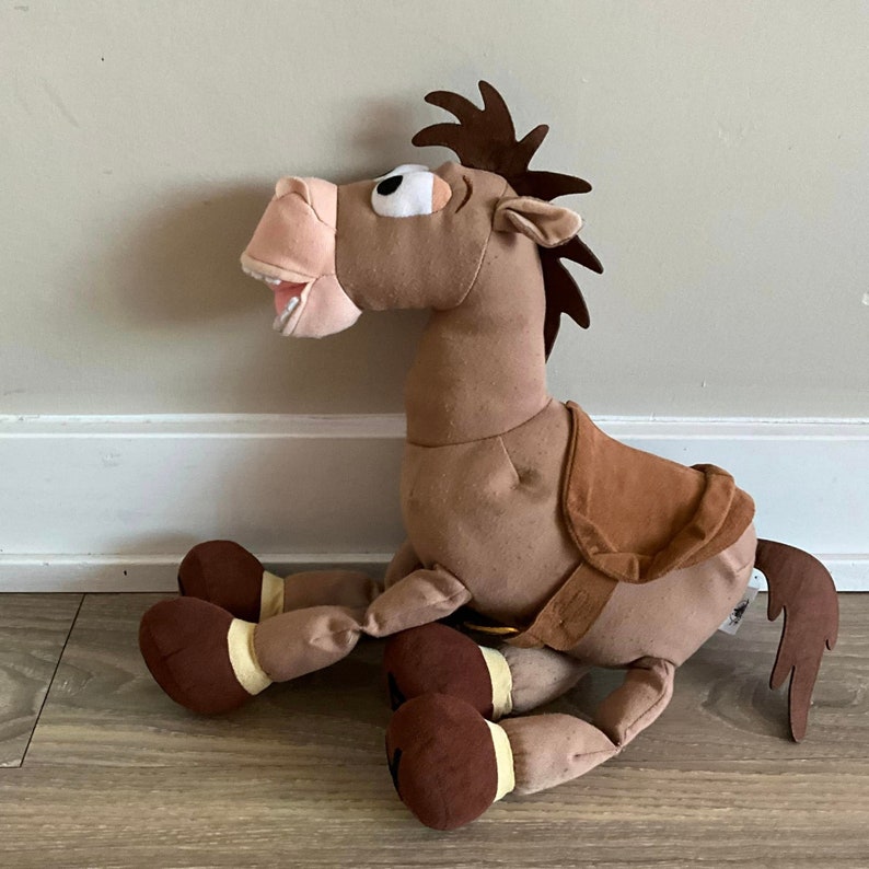 Puede incluir: Un caballo de peluche marr&oacute;n con una silla de montar, ojos marrones y una boca muy abierta. El caballo est&aacute; sentado en un suelo de madera.