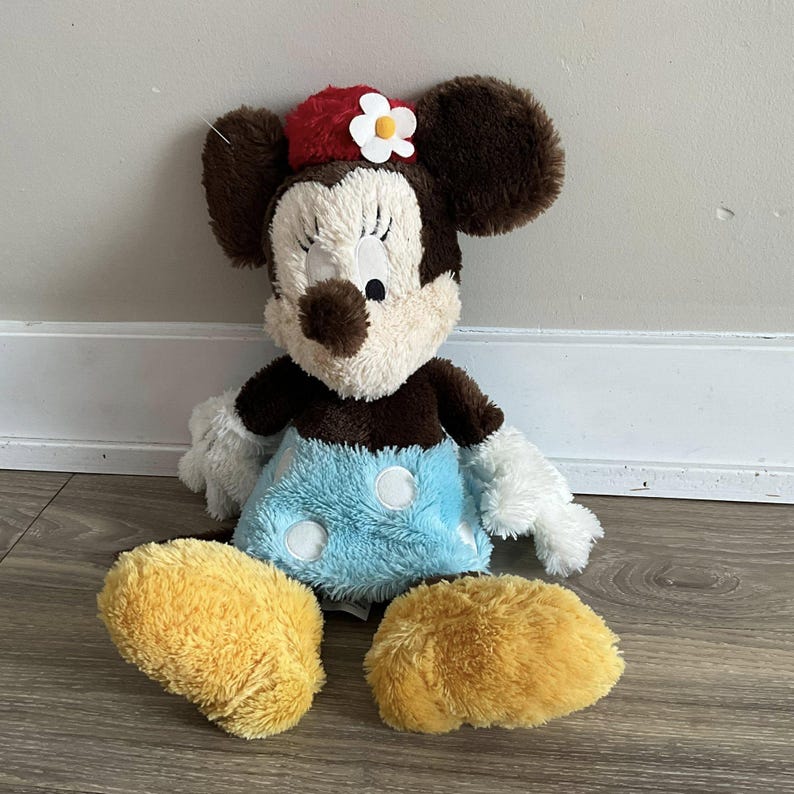 May include: A plush Minnie Mouse doll with brown ears, a red hat with a white flower, and a light blue skirt with white polka dots. The doll has white gloves and yellow shoes. The doll is sitting on a wooden floor.