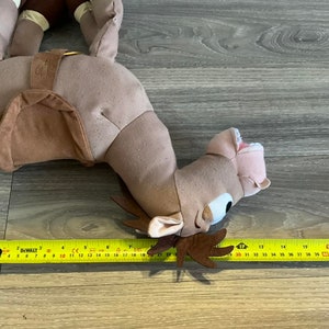 Puede incluir: Un camello de peluche marr&oacute;n con una silla de montar y una correa de cuero marr&oacute;n. El camello tiene una boca rosa y ojos grandes azules. El camello est&aacute; acostado de lado con la cabeza girada hacia la derecha. El camello mide aproximadamente 56 cm de largo.
