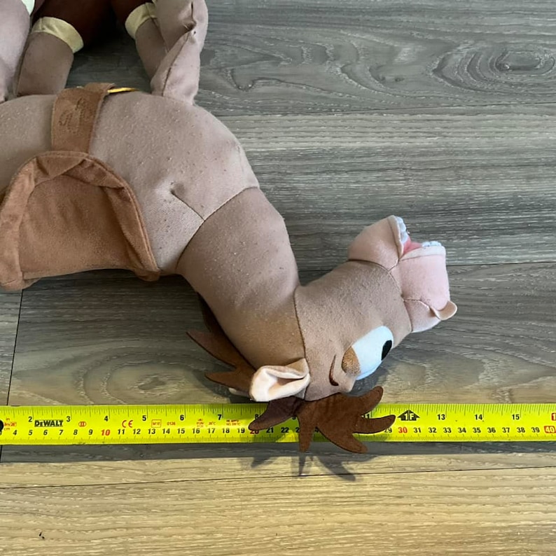 Puede incluir: Un camello de peluche marr&oacute;n con una silla de montar y una correa de cuero marr&oacute;n. El camello tiene una boca rosa y ojos grandes azules. El camello est&aacute; acostado de lado con la cabeza girada hacia la derecha. El camello mide aproximadamente 56 cm de largo.