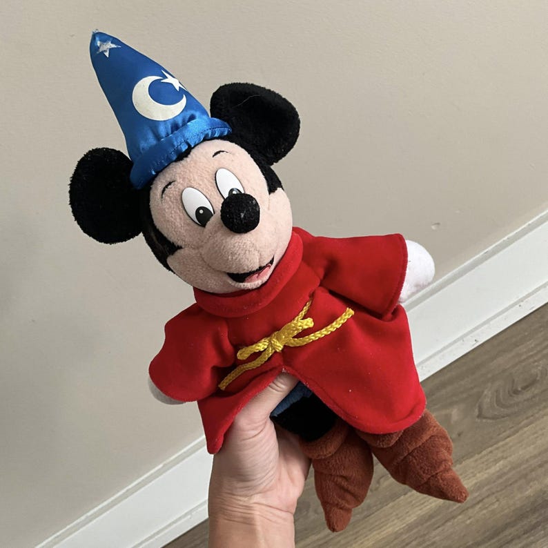 May include: A plush Mickey Mouse toy dressed as a sorcerer. Mickey wears a blue wizard hat with a crescent moon and star, a red robe with a gold rope tie, and brown boots. The toy has black ears and a smiling face.