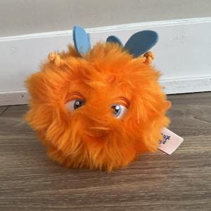 May include: A plush, orange, fuzzy creature with blue wing-like appendages and large, expressive eyes. The toy has a tag attached. The toy is designed to look like a cartoon character.