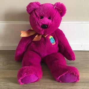May include: A plush purple teddy bear with a gold ribbon around its neck. The bear has a small embroidered patch on its chest that says "2000".
