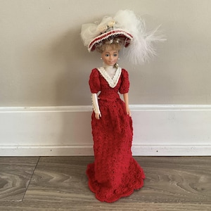 May include: A doll in a red crocheted Victorian-style gown with a white lace collar and cuffs. The doll has a matching hat with red and white trim, and white feathers. The doll is standing on a wooden floor.