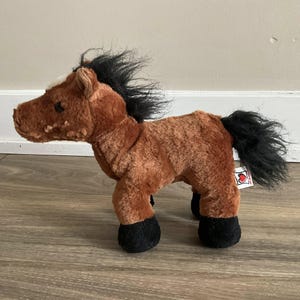 Op de afbeelding: Een bruin pluche paard met een zwarte manen en staart. Het paard heeft zwarte hoeven en een witte vlek op het voorhoofd. Een klein label met een hartje is aan het paard bevestigd.