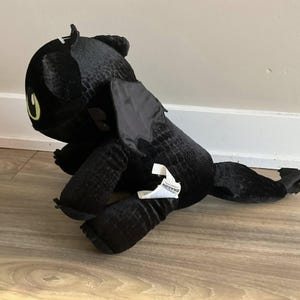 Può includere: Un peluche di drago nero con grandi occhi gialli e un corpo strutturato. Il drago ha grandi ali e un'etichetta attaccata alla zampa. Il giocattolo è appoggiato su un pavimento di legno.