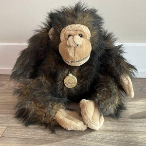 May include: A soft chimpanzee toy with brown and black fur, tan face, hands, and feet. The stuffed animal has a gold-coloured medallion hanging from its neck. The toy is sitting on a wooden surface.