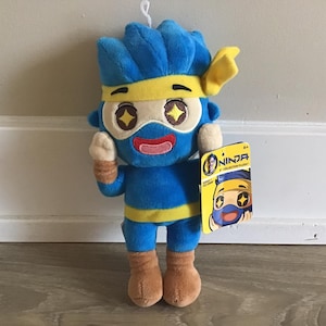 May include: Blue and yellow plush toy of a cartoon ninja character with large eyes and a yellow headband. The toy is holding its right hand up in a wave. The toy is standing on a wooden floor. The toy has a tag that says "Ninja Collector Plush".