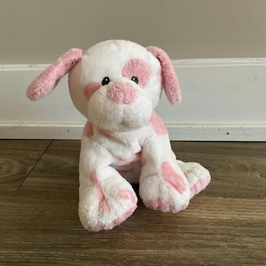Puede incluir: Un peluche de perro en posición sentada. El juguete es blanco con manchas rosas en el cuerpo, las orejas y las patas. Tiene ojos negros y una nariz rosa. El juguete está hecho de material suave y difuso.
