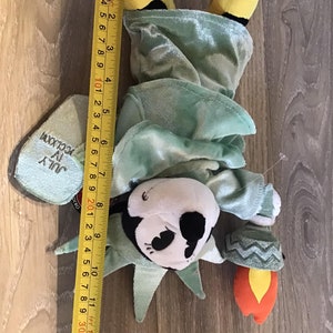 May include: A plush toy of the Statue of Liberty, dressed in a green robe with a yellow crown. The toy is holding a torch in its right hand. The toy is approximately 11 inches tall.