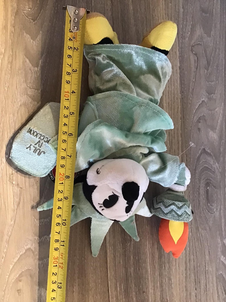 May include: A plush toy of the Statue of Liberty, dressed in a green robe with a yellow crown. The toy is holding a torch in its right hand. The toy is approximately 11 inches tall.
