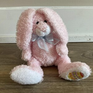 May include: A pink plush bunny with long, floppy ears and a light blue ribbon. The bunny has white accents on its feet and a small embroidered design. The toy is sitting on a wooden surface.