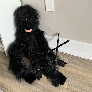 May include: A plush, black gorilla puppet with a smiling face and long, shaggy fur. The puppet has black hands and feet, and is controlled by black sticks and strings. The puppet is sitting on a wooden floor.