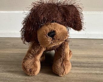 Op de afbeelding: Een zacht, bruin pluche hondenspeelgoed met een harige kop en een zacht lichaam. De hond heeft een rond, lichtbruin gezicht met een zwarte neus. Het speelgoed staat op zijn vier poten. Het speelgoed is ontworpen om op een hond te lijken.