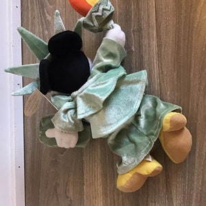 May include: A plush toy of Mickey Mouse dressed as the Statue of Liberty. The toy is green and brown with a black head and a torch in its hand.
