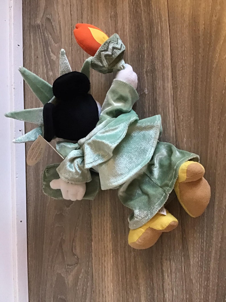 May include: A plush toy of Mickey Mouse dressed as the Statue of Liberty. The toy is green and brown with a black head and a torch in its hand.
