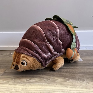 Könnte beinhalten: Ein Plüschtier-Gürteltier mit braunem Panzer und beigem Körper. Das Spielzeug hat ein freundliches Gesicht mit großen Augen und ein kleines orangefarbenes und grünes Accessoire auf dem Rücken. Das Gürteltier liegt auf einer Holzoberfläche.