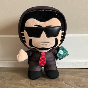 May include: A plush toy character wearing a dark brown hooded jacket, black sunglasses, and a red tie with a crown pattern. The character has black hair, sideburns, and is holding a small fan of green paper money bills labeled with 100 and a dollar sign.