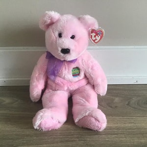 May include: Pink Ty Beanie Baby plush bear with a purple ribbon and a green, yellow and red embroidered patch on its chest. The bear has a black nose and black eyes. The tag reads "Ty".