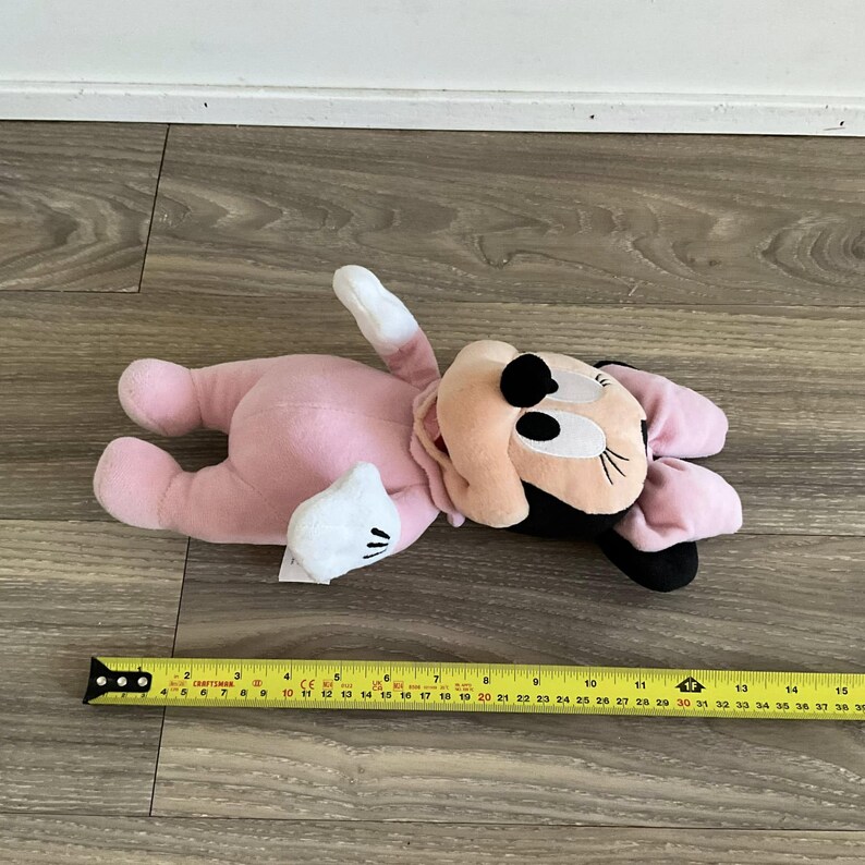 Pu&ograve; includere: Un peluche rosa di Minnie Mouse sdraiato su una superficie di legno. Il peluche ha un corpo rosa, un viso beige, occhi neri e un fiocco nero. Il giocattolo &egrave; lungo circa 38 cm. Un metro a nastro giallo &egrave; accanto al giocattolo.