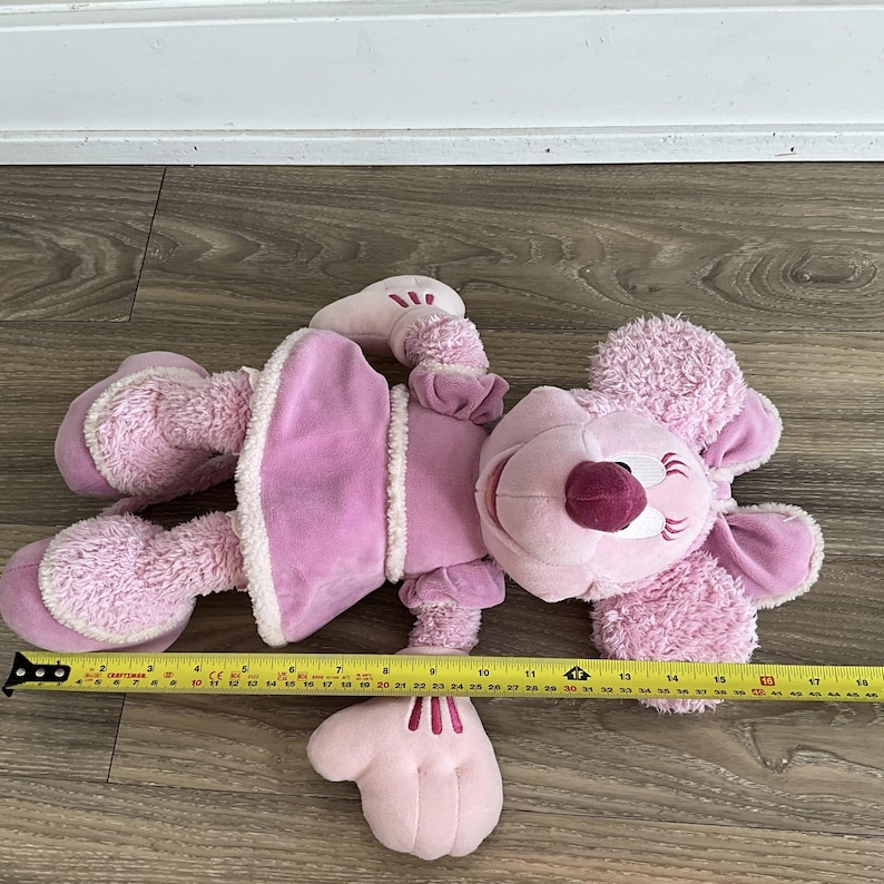 Peut inclure: Une peluche rose avec un visage souriant, de grandes oreilles et une robe. Le jouet mesure environ 46 cm de long, mesur&eacute; avec un m&egrave;tre ruban jaune. Le jouet a un corps rose clair avec des accents roses plus fonc&eacute;s.
