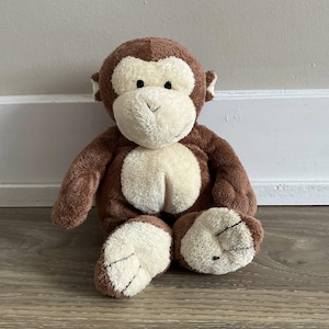 以下が含まれることがあります： 茶色の毛皮とクリーム色の顔、手、足を持つぬいぐるみのおもちゃの猿。 ぬいぐるみは、足を伸ばし、腕を横に置いて直立しています。 猿はフレンドリーな表情をしています。