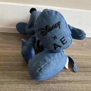 K&ouml;nnte beinhalten: Ein blaues Denim-Pl&uuml;schtier mit schwarzem, gesticktem Text "Disney x AE". Das Spielzeug hat eine schwarze Knopf-Nase und liegt auf einem Holzboden. Das Spielzeug hat eine hellblaue Farbe.