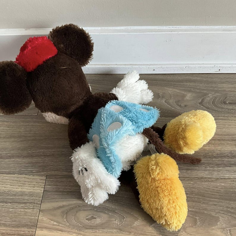 May include: A plush Mickey Mouse toy in a lying position. The toy has a brown body, large black ears, and a red hat. It has white gloves, a blue and white patterned shirt, and yellow feet. The toy is lying on a wooden surface.