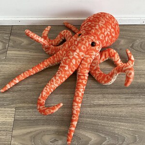 May include: A plush octopus toy in shades of orange and white. The octopus has a round head, black eyes, and eight long, patterned tentacles. The toy is made of soft fabric and is designed to be cuddly.