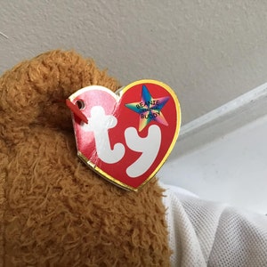 May include: Close-up of a brown and tan plush toy with a red and white Ty Beanie Original Buddy tag attached.