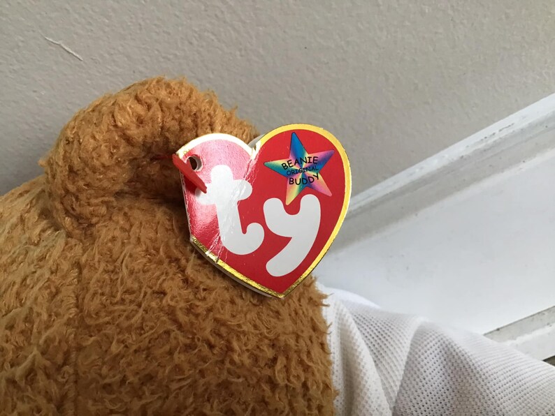 May include: Close-up of a brown and tan plush toy with a red and white Ty Beanie Original Buddy tag attached.