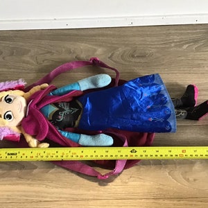 May include: A plush Frozen backpack featuring a pink and purple hooded character with blonde hair and a blue dress. The backpack has a zipper closure and adjustable straps.