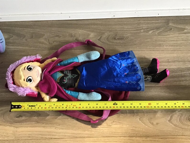 May include: A plush Frozen backpack featuring a pink and purple hooded character with blonde hair and a blue dress. The backpack has a zipper closure and adjustable straps.