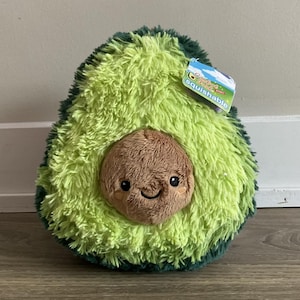Puede incluir: Un juguete de peluche con forma de aguacate con una cara sonriente y un hueso marrón. El aguacate es verde con un borde verde más oscuro. Una etiqueta dice "Squishable".
