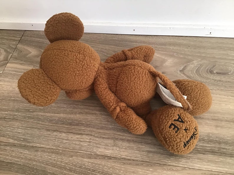 Puede incluir: Un juguete de peluche marr&oacute;n con forma de Mickey Mouse. El juguete est&aacute; tumbado de lado con la cabeza apoyada en el suelo. El juguete tiene el texto "A Bathing Ape x Disney" en su pie.